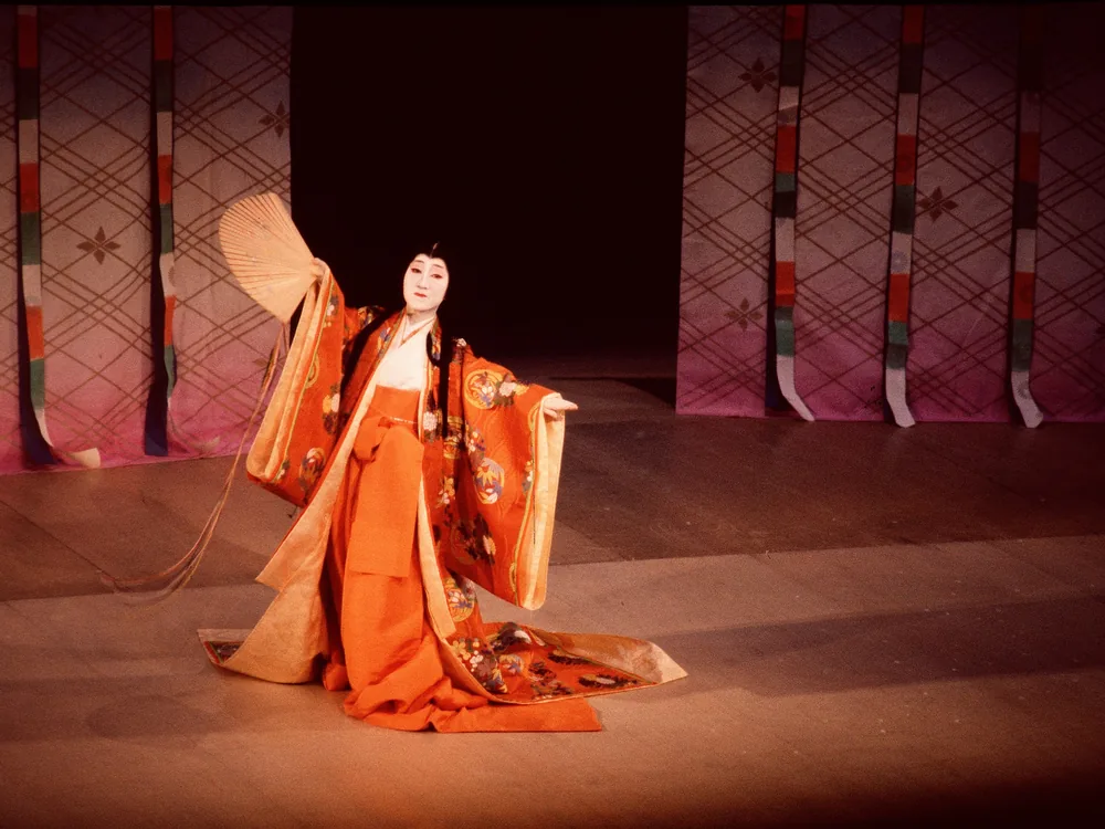 A dancer in an orange layered kimono holding an open fan on a Kyoto stage in the Gion area