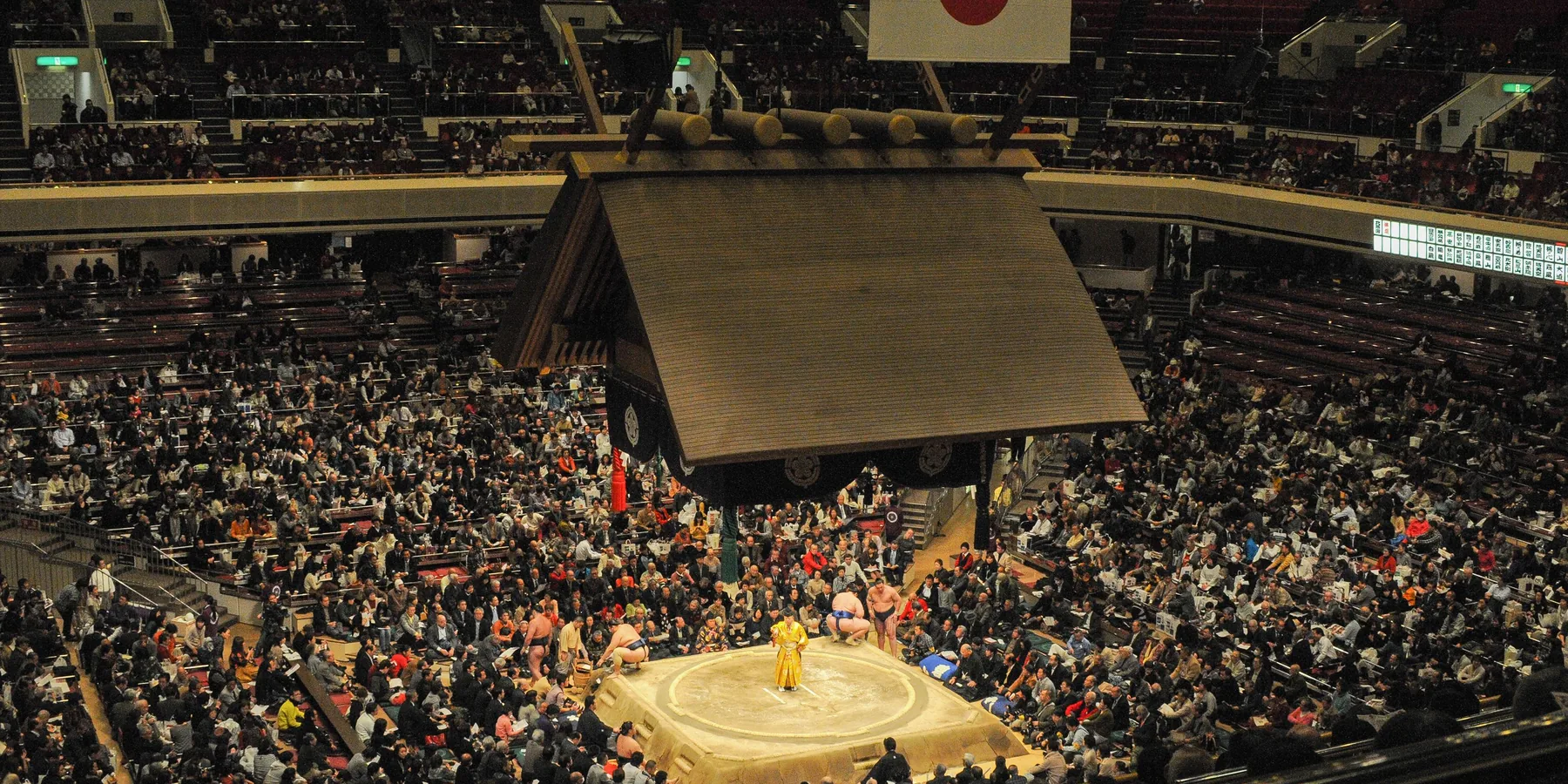 A packed sumo arena in Japan seen from above, with a hanging shrine-style roof over the raised ring